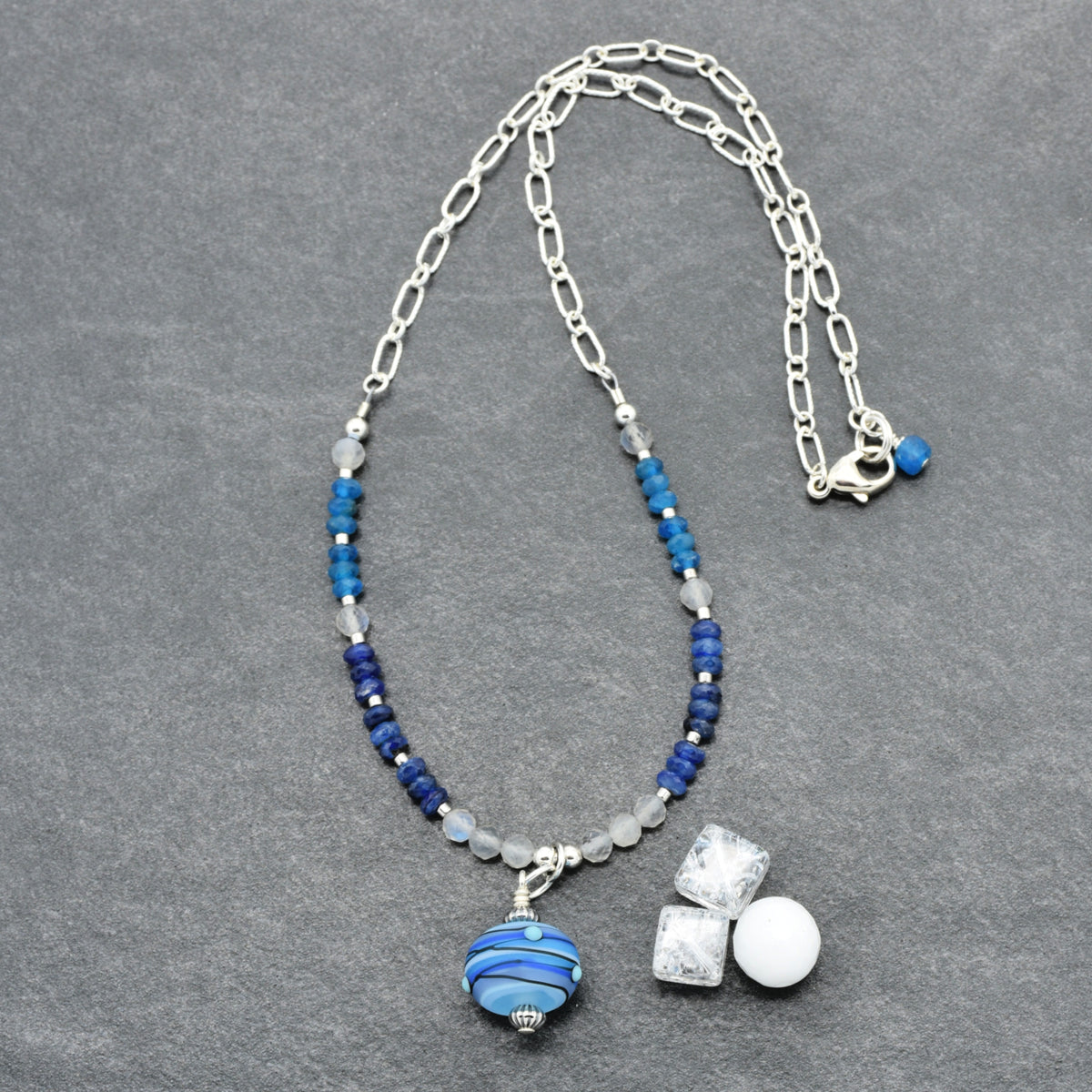 A silver chain necklace with blue beads, a blue and white striped glass bead pendant, displayed on a textured gray background.