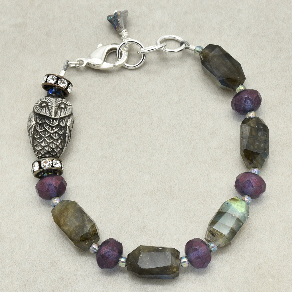 A beaded bracelet featuring faceted purple and smoky gray beads, a silver-toned owl charm, and a lobster clasp, displayed on a neutral beige background.