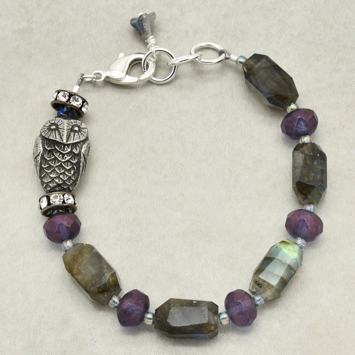 A beaded bracelet featuring faceted purple and smoky gray beads, a silver-toned owl charm, and a lobster clasp, displayed on a neutral beige background.