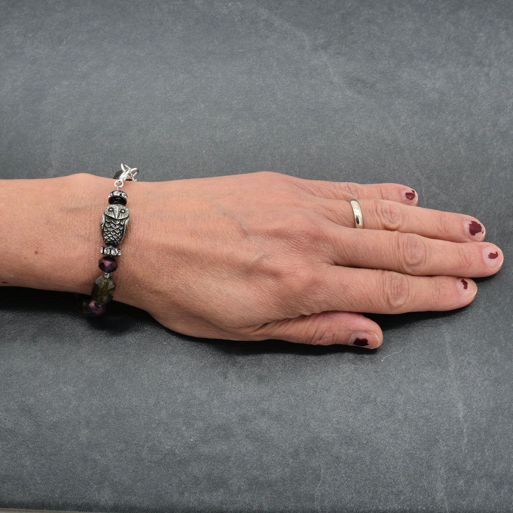 A silver-toned beaded bracelet with an owl charm and purple beads, shown on a person's wrist against a dark gray textured background.
