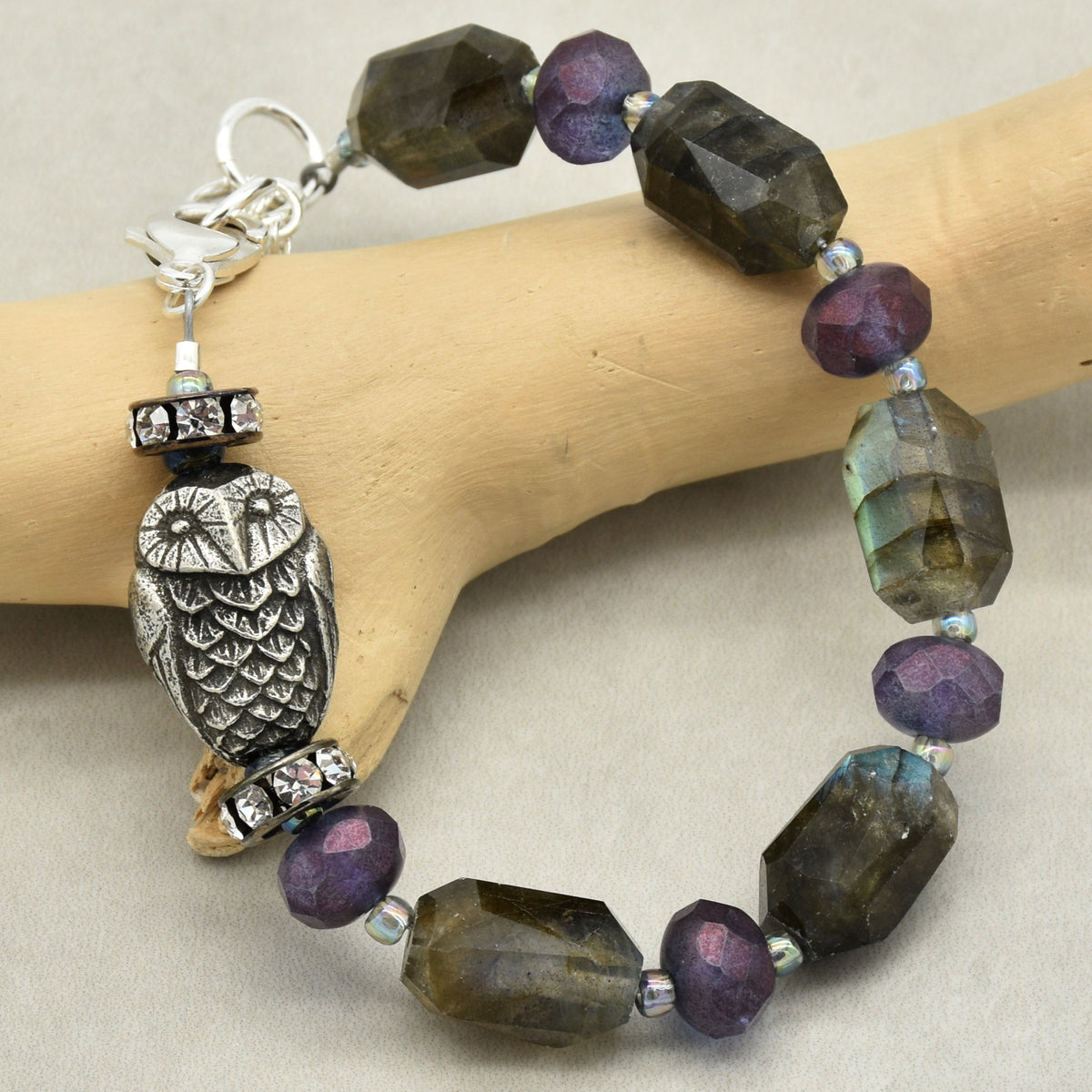 A beaded bracelet with faceted dark gray and purple beads, a silver-toned owl charm, and rhinestone accents, displayed on a light wood stand against a neutral background.