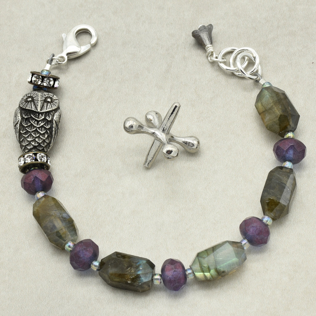 A silver beaded bracelet featuring faceted labradorite beads, purple glass rondelle beads, and a detailed owl charm, displayed on a neutral beige background.