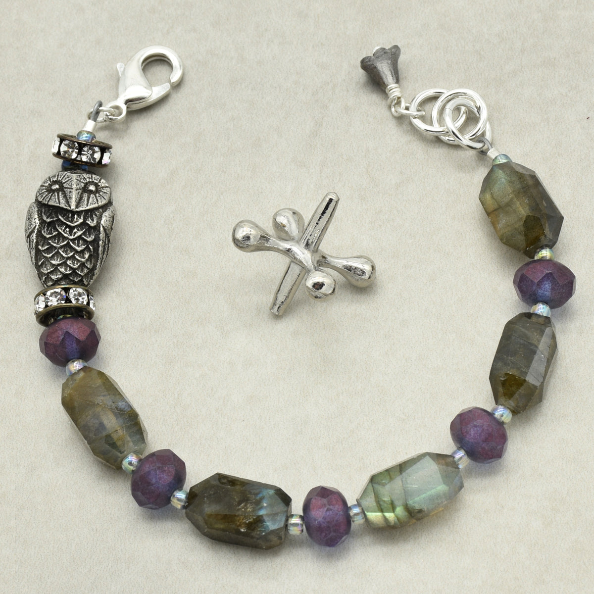 A silver beaded bracelet featuring faceted labradorite beads, purple glass rondelle beads, and a detailed owl charm, displayed on a neutral beige background.