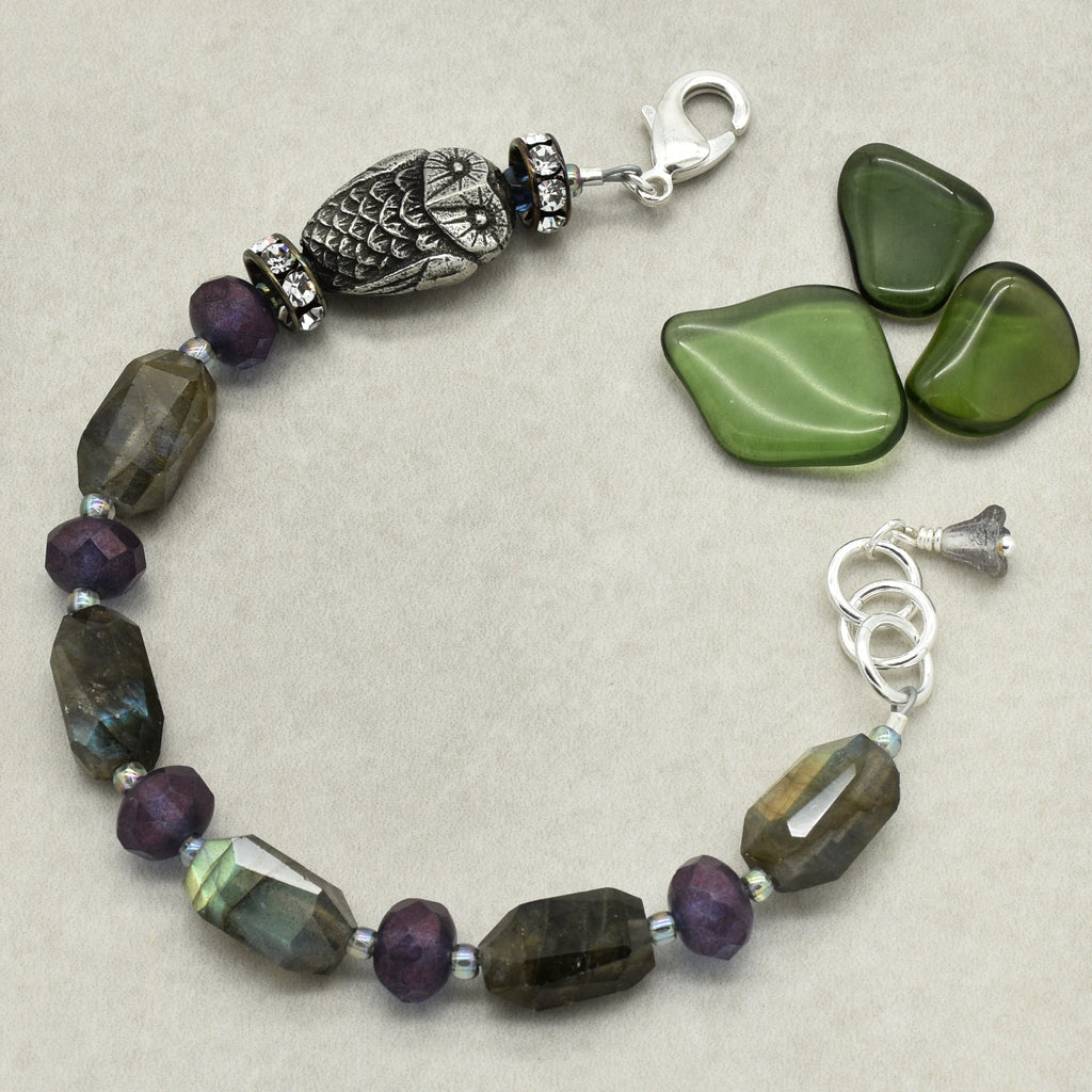 A beaded bracelet with faceted green, purple, and iridescent beads, a silver owl charm, and three green stone pieces as props on a neutral beige background.