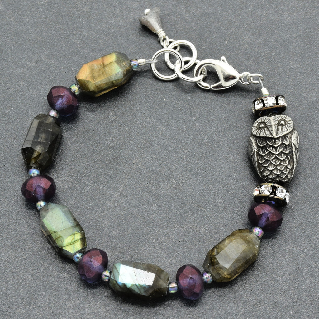 A beaded bracelet featuring faceted labradorite beads with iridescent green and gold flashes, dark plum faceted beads, a silver-toned owl charm, and a silver lobster clasp, displayed on a dark gray textured background.