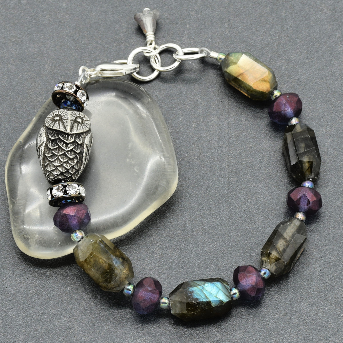 A beaded bracelet with a silver-toned owl charm, faceted labradorite beads, and iridescent purple beads, displayed on a dark gray background next to a clear glass stone.