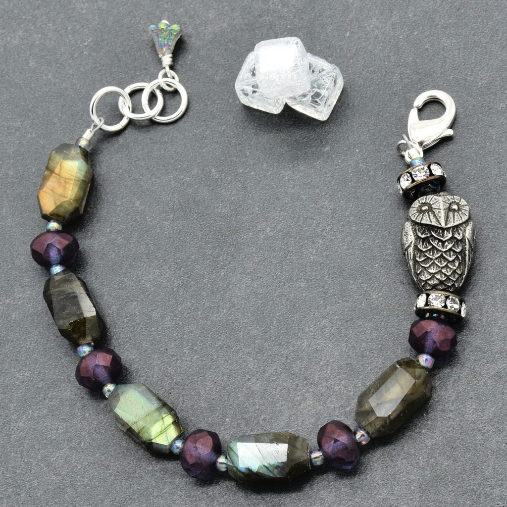 A beaded bracelet featuring faceted labradorite beads, dark purple beads, a silver owl charm, and a silver lobster clasp, displayed on a dark gray background with two raw crystal clusters.