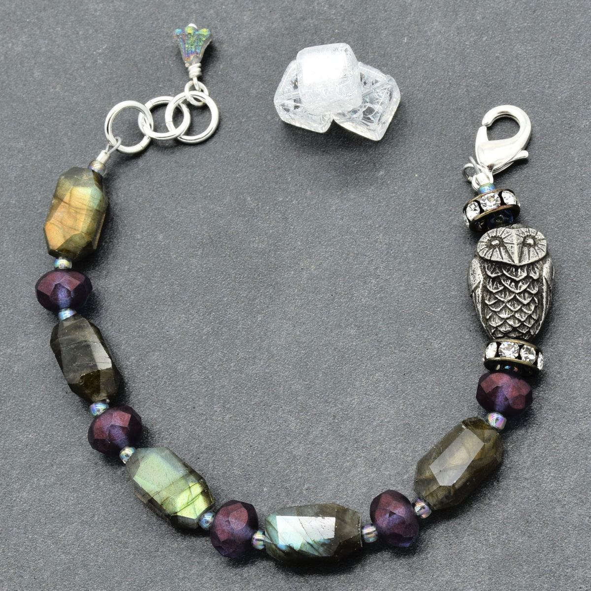 A beaded bracelet featuring faceted labradorite beads, dark purple beads, a silver owl charm, and a silver lobster clasp, displayed on a dark gray background with two raw crystal clusters.