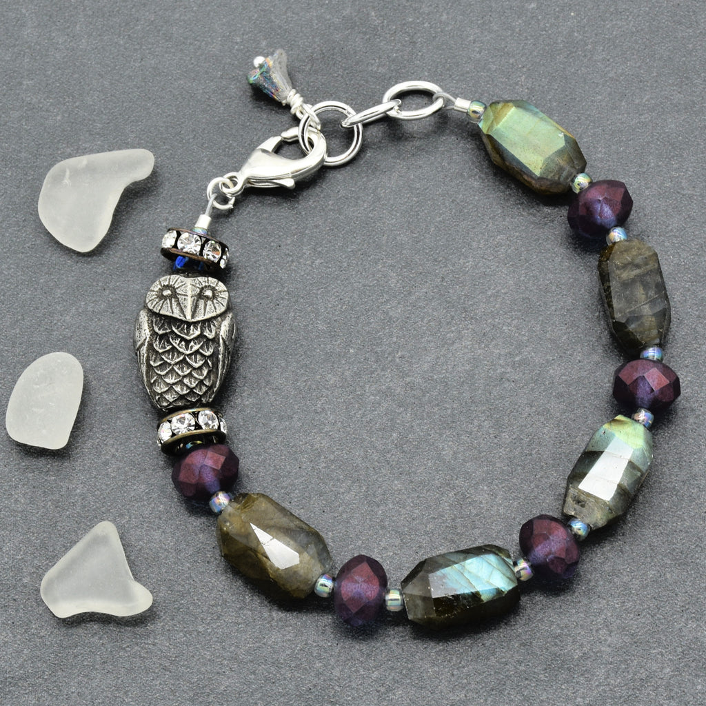 A beaded bracelet featuring a central silver owl charm, faceted labradorite beads showing green and blue iridescence, deep purple beads, and small blue accent beads, displayed on a dark gray background with three clear glass pebble props.