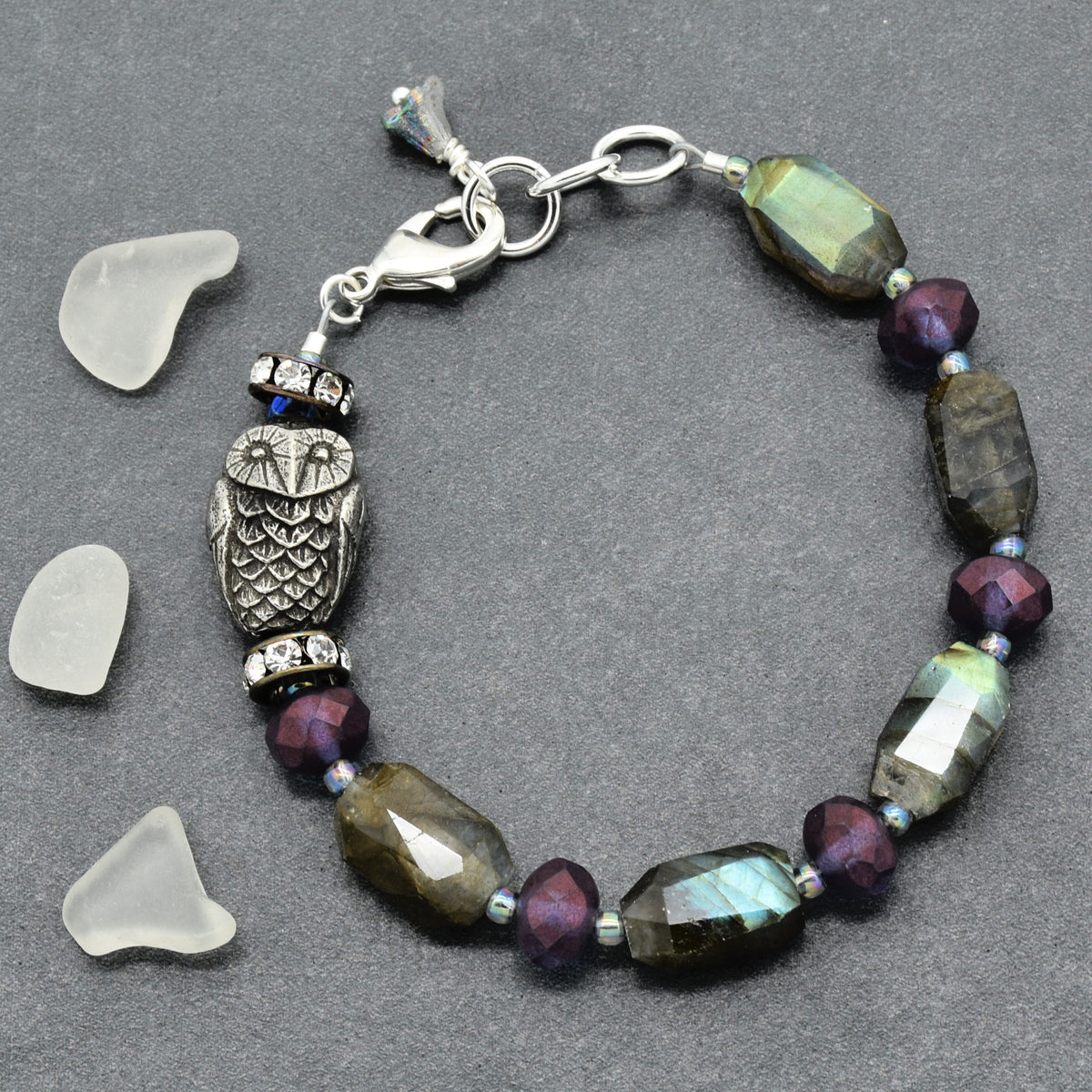A beaded bracelet featuring a central silver owl charm, faceted labradorite beads showing green and blue iridescence, deep purple beads, and small blue accent beads, displayed on a dark gray background with three clear glass pebble props.