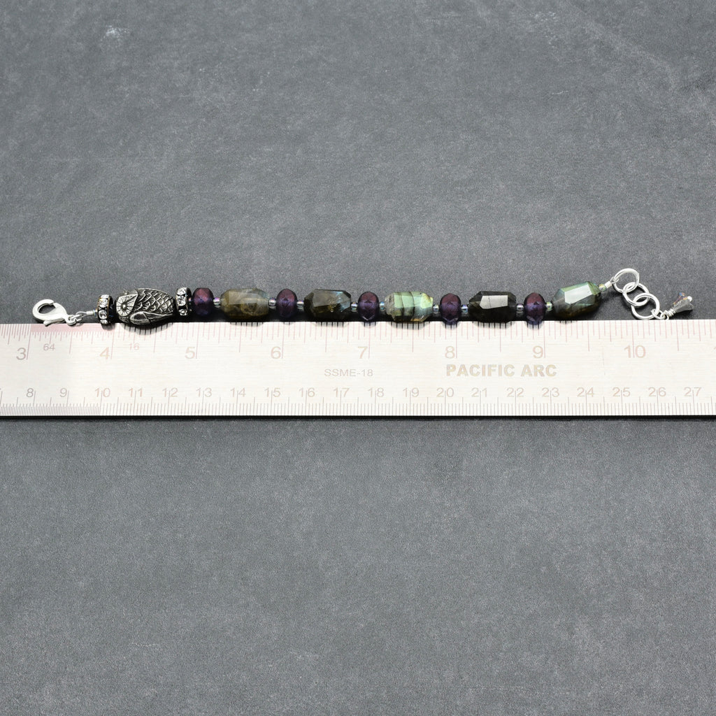 A beaded bracelet with faceted labradorite beads, featuring a silver owl charm and silver clasp, shown next to a ruler for scale on a gray textured background.