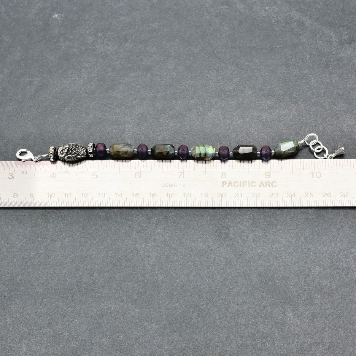 A beaded bracelet with faceted labradorite beads, featuring a silver owl charm and silver clasp, shown next to a ruler for scale on a gray textured background.