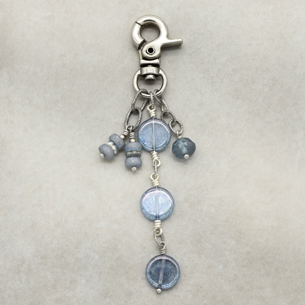 A silver-toned lobster clasp keychain or bag charm featuring three dangling beaded strands with round light blue beads and small metallic spacer beads, displayed on a neutral beige textured background.