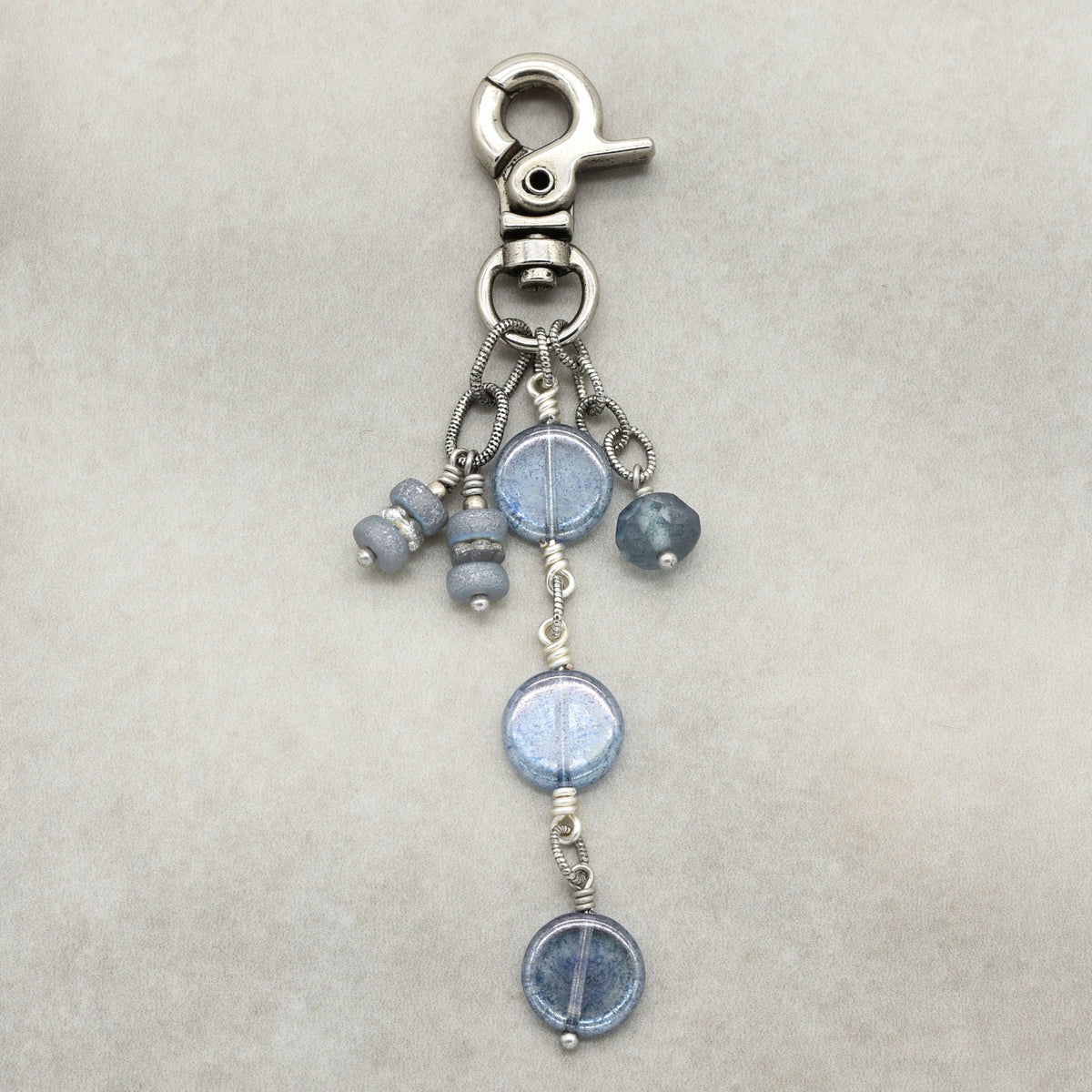 A silver-toned lobster clasp keychain or bag charm featuring three dangling beaded strands with round light blue beads and small metallic spacer beads, displayed on a neutral beige textured background.