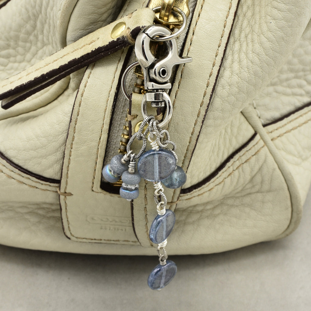 Close-up of a handbag in light beige pebbled leather, featuring a silver-tone lobster clasp and charm with blue faceted beads.