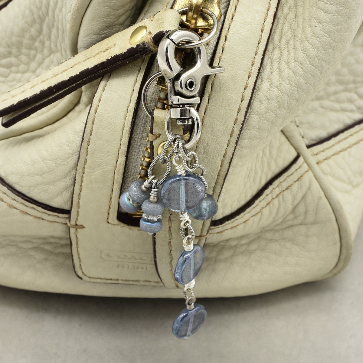 Close-up of a handbag in light beige pebbled leather, featuring a silver-tone lobster clasp and charm with blue faceted beads.