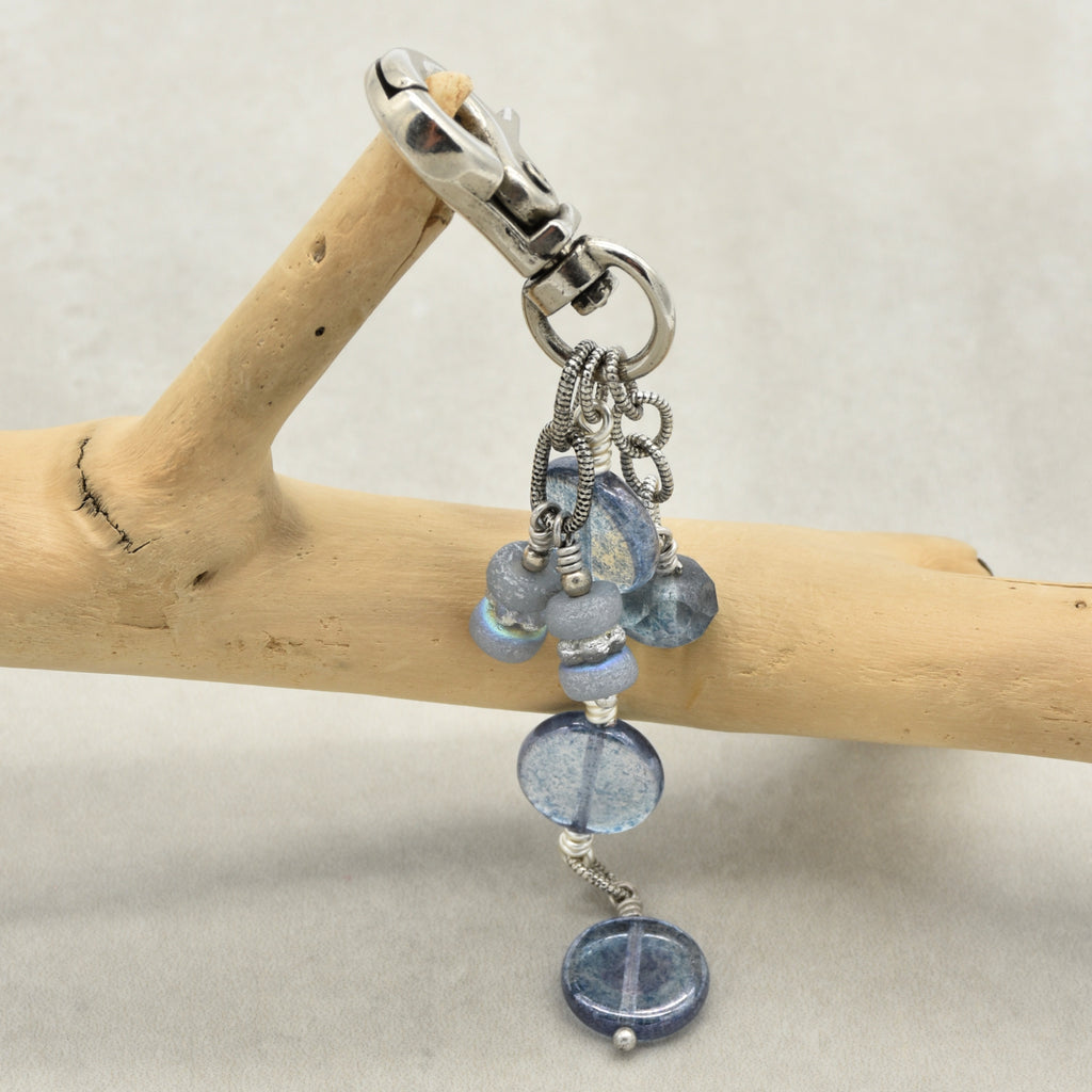 A beaded keychain charm with light blue glass beads, silver wire wrapping, and a silver lobster clasp, hanging from a light wood branch against a neutral gray background.