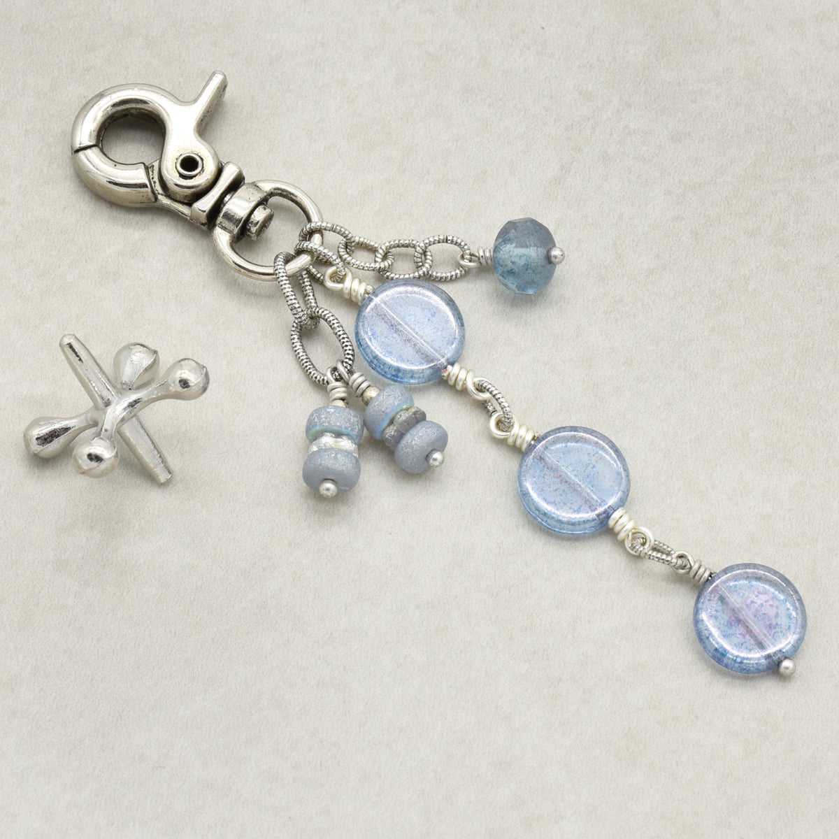 A silver-toned lobster clasp keychain or bag charm with dangling beaded chains. The beaded strands feature faceted light blue beads and smaller gray-blue spacer beads, all displayed on a neutral beige textured background.