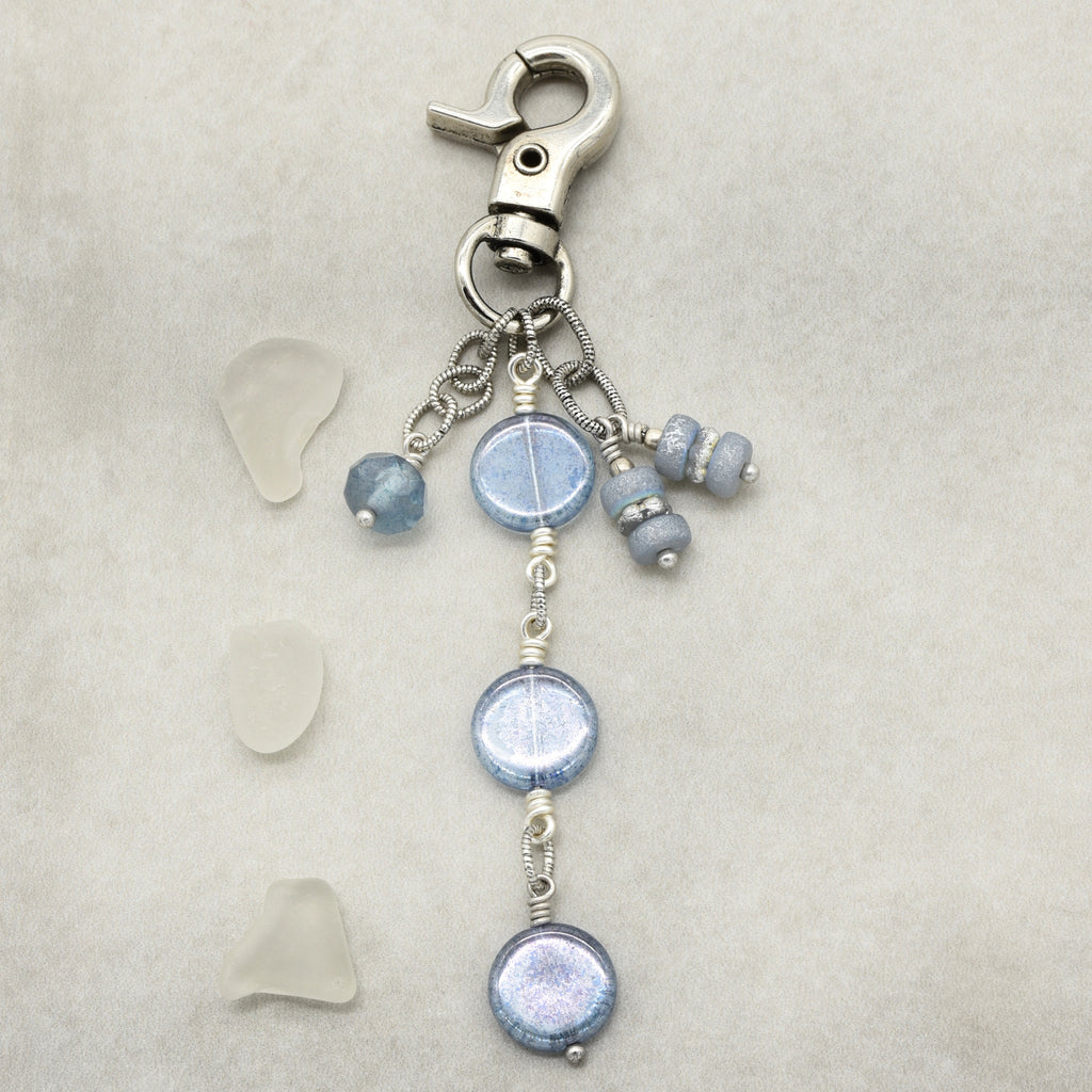 A handmade beaded keychain or bag charm featuring a silver lobster clasp, a central round iridescent blue bead, three smaller blue beads, and three dangling wire-wrapped beads, displayed on a textured light beige background with three small pieces of sea glass to the left.
