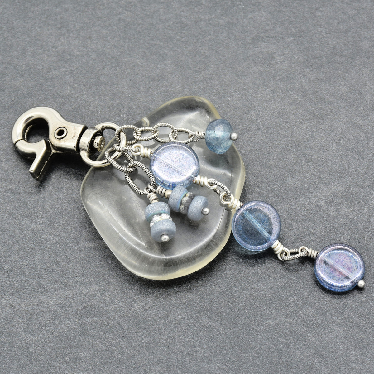 A handmade keychain featuring a polished silver-tone lobster clasp, and dangling blue glass beads and chains on a textured gray background.