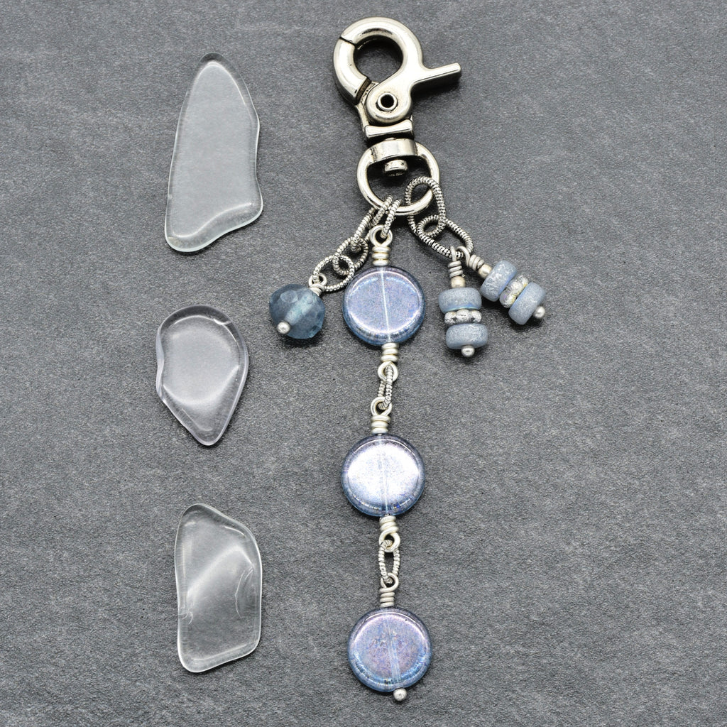 A handmade beaded keychain or bag charm with a silver lobster clasp, featuring three blue-gray glass beads and three small wrapped beads, displayed alongside three clear glass chips on a dark gray slate background.