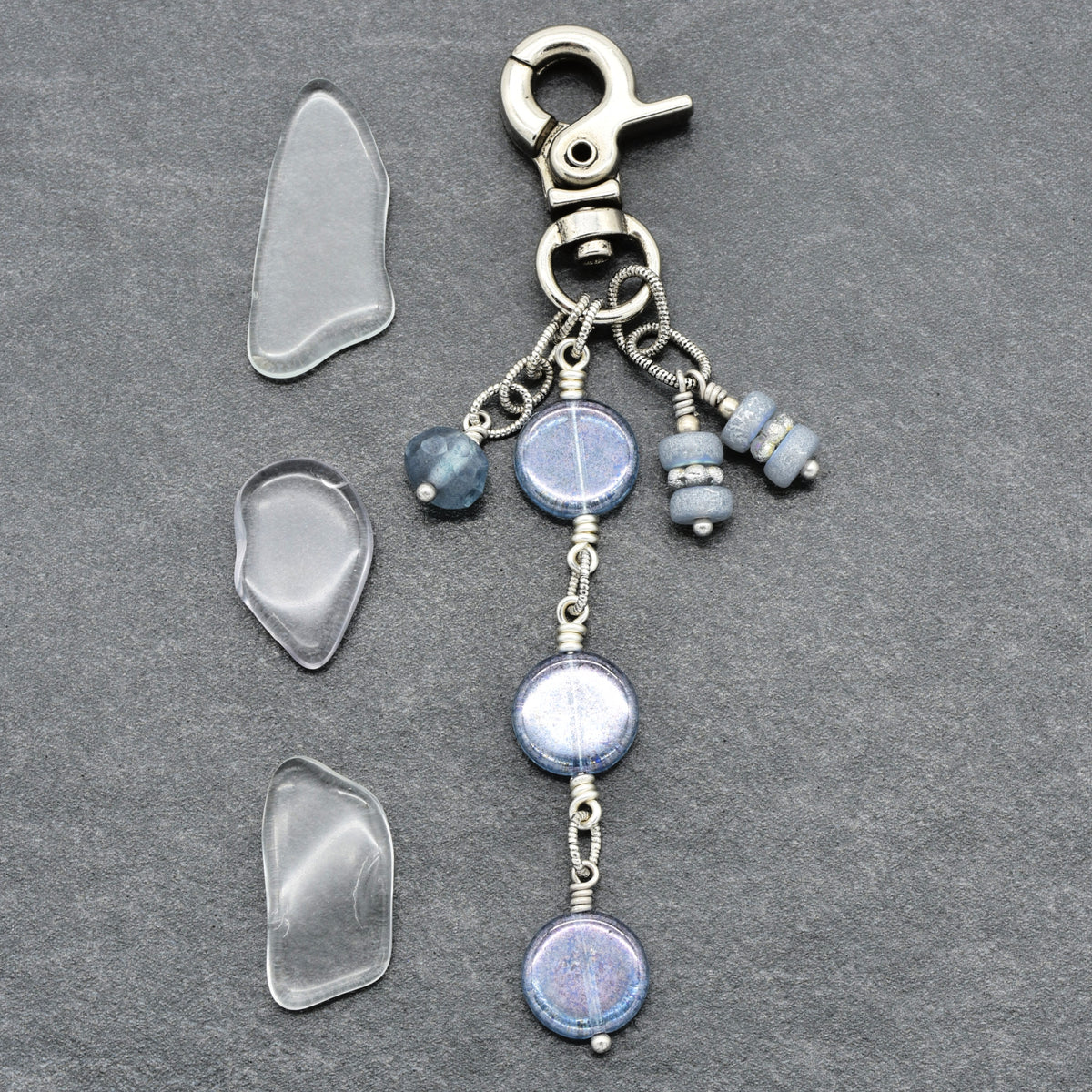 A handmade beaded keychain or bag charm with a silver lobster clasp, featuring three blue-gray glass beads and three small wrapped beads, displayed alongside three clear glass chips on a dark gray slate background.