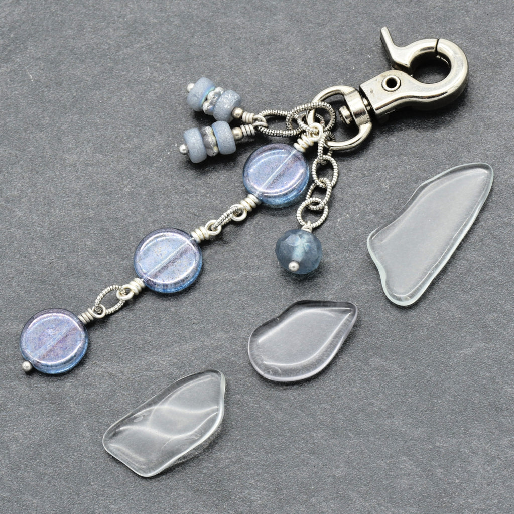 A handmade keychain or bag charm with a silver-toned lobster clasp, featuring light blue and white marbled glass beads, and a small blue bead, on a dark gray textured background.