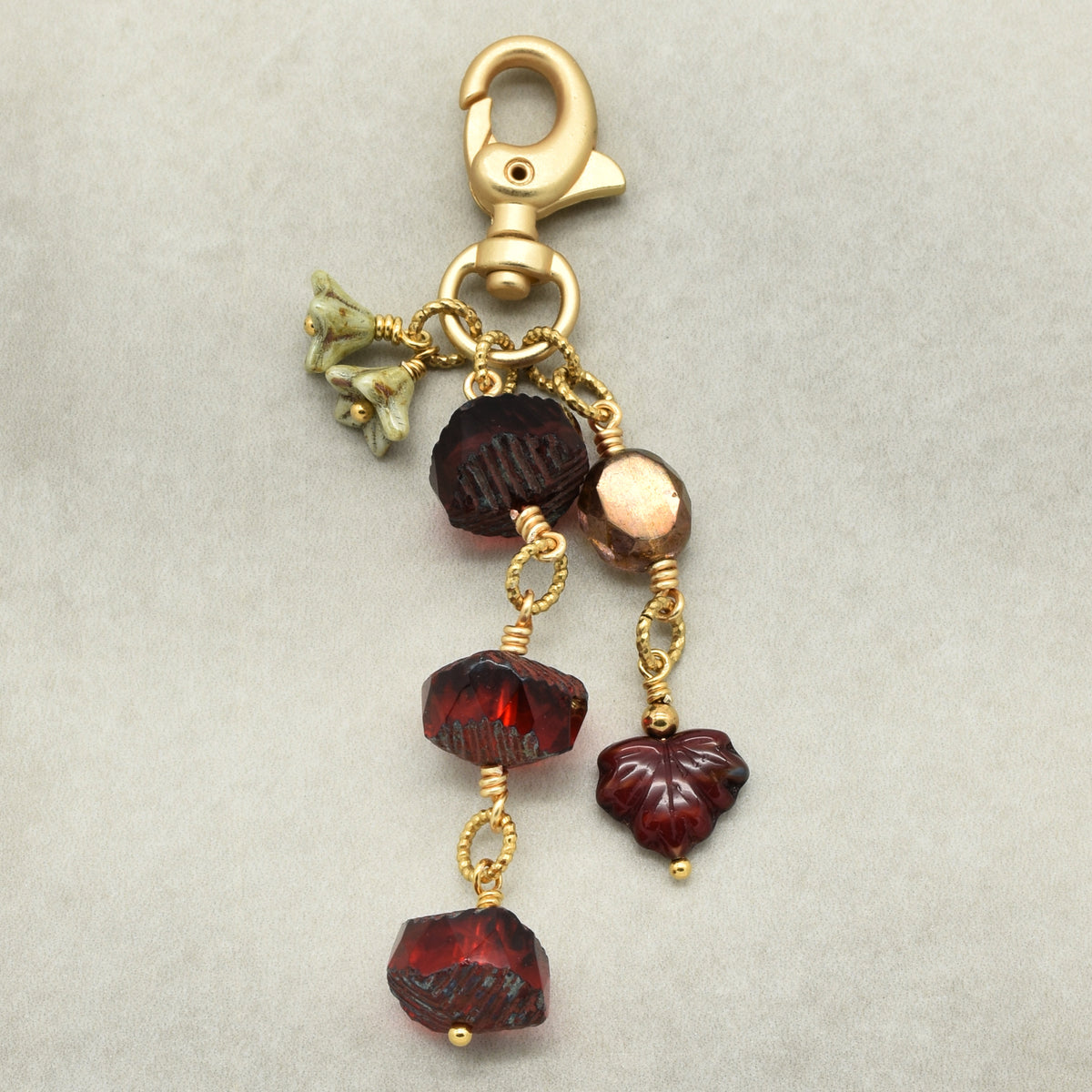 A gold-toned lobster clasp keychain or bag charm featuring dark red faceted beads, a small metallic rose gold bead, green bellflower charms, and a carved dark red maple leaf bead, displayed on a neutral gray background.