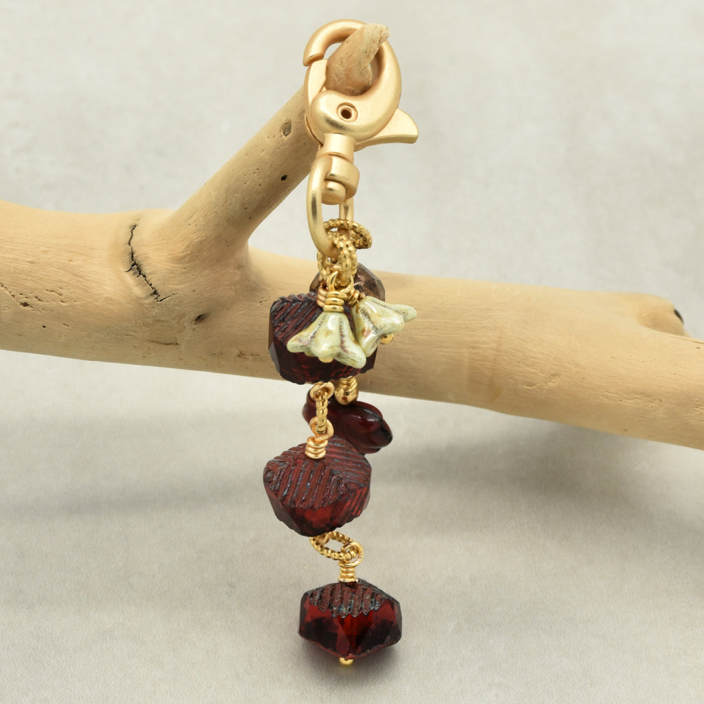 A handmade dangle handbag charm with faceted dark red beads, a small light green bellflower charms, and gold-toned metal hardware, hanging from a light wood branch against a neutral gray background.