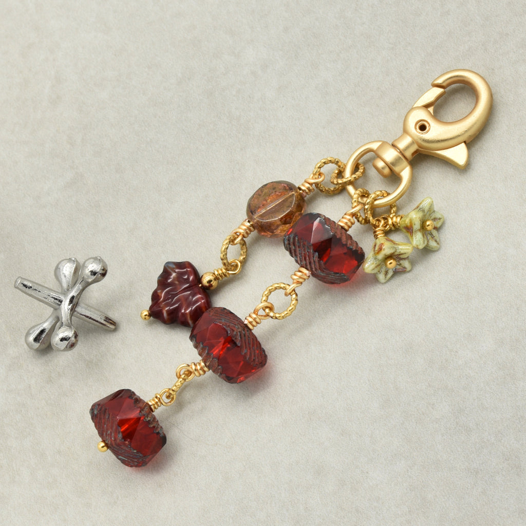 A gold-toned beaded keychain or bag charm with faceted red and amber glass beads, small green floral charms, and a lobster clasp, shown on a light gray background with a separate silver jack beside it.