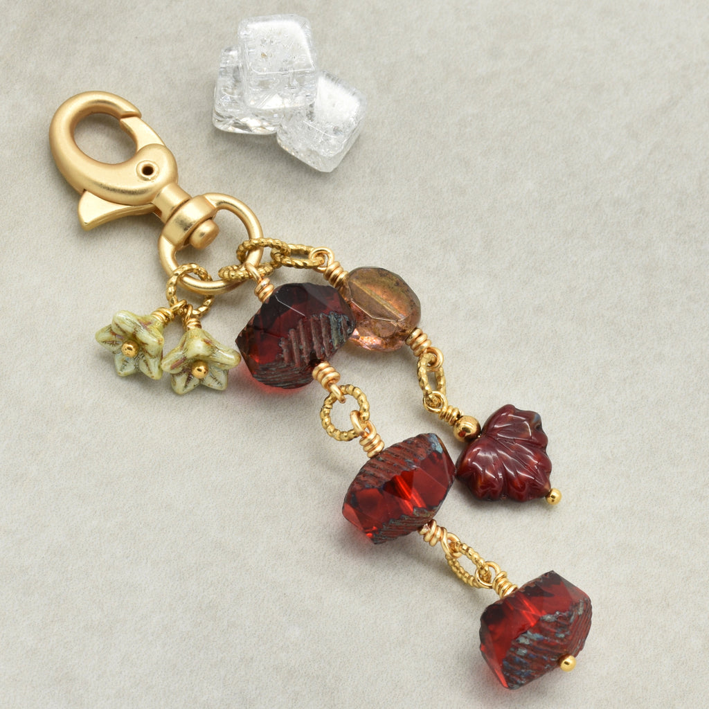 A gold-toned lobster clasp keychain or bag charm with dangling faceted beads in deep red, amber-brown, and small pale green flower charms, photographed on a light gray background.