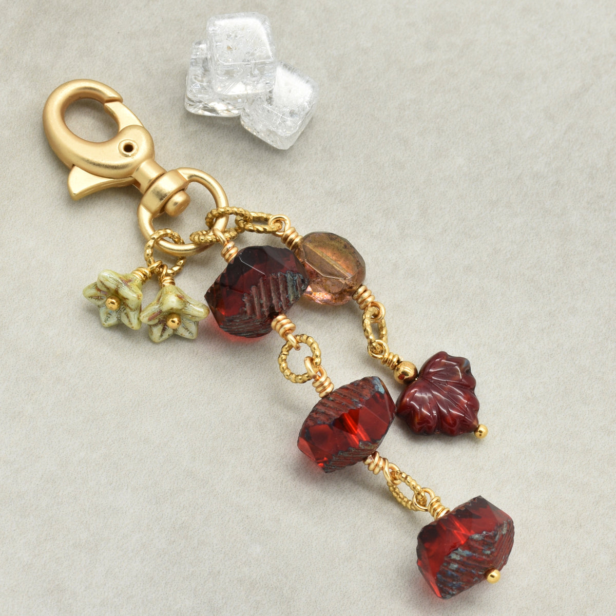 A gold-toned lobster clasp keychain or bag charm with dangling faceted beads in deep red, amber-brown, and small pale green flower charms, photographed on a light gray background.