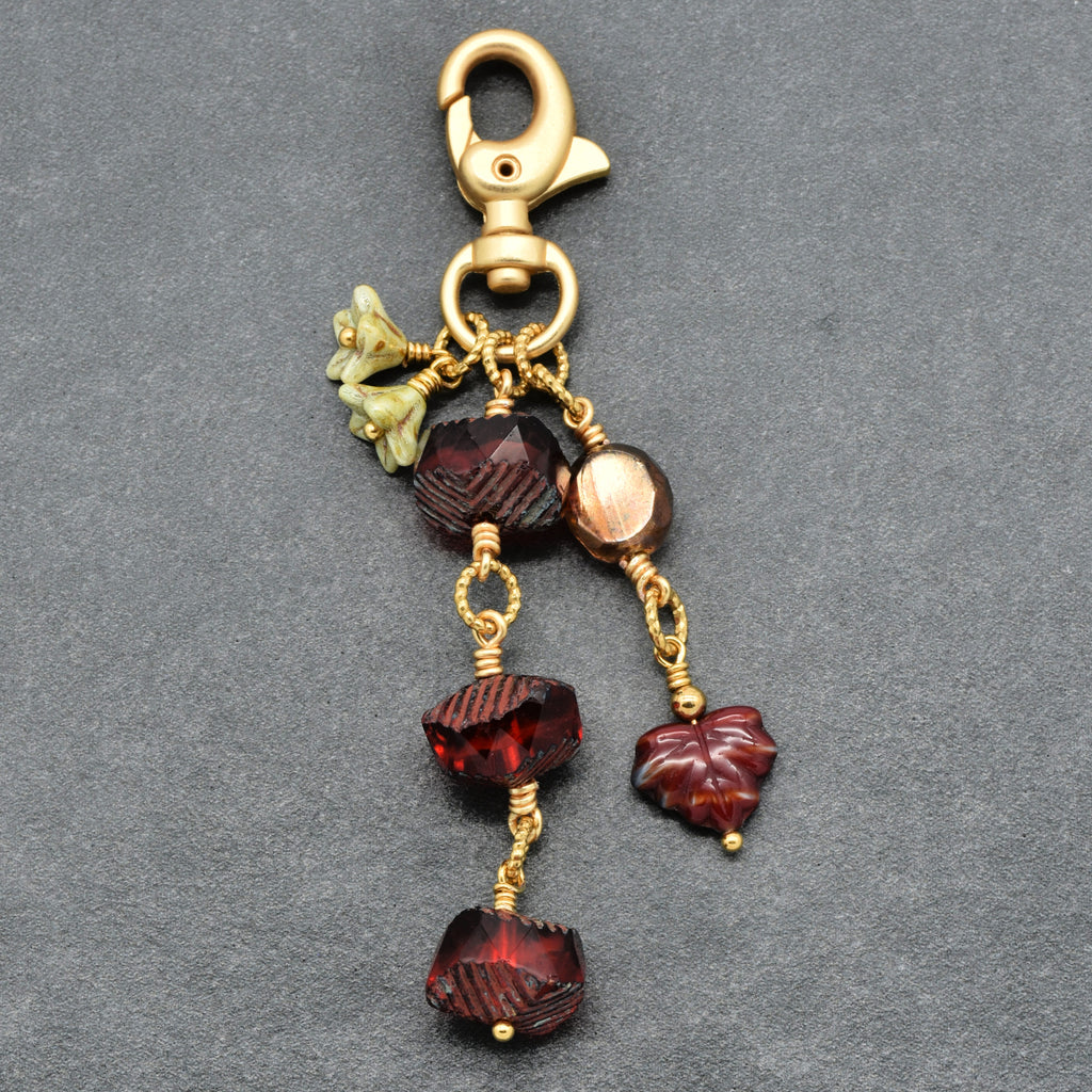 A gold-tone metal keychain or bag charm with a lobster clasp, featuring carved dark red glass beads, small pale green bellflower charms, and a maple leaf-shaped red bead, displayed on a textured gray background.