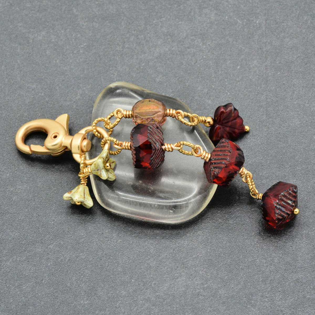 A gold-tone keychain or bag charm with a lobster clasp, featuring faceted dark red beads, and a small green leaf charm, displayed on a dark gray textured background.