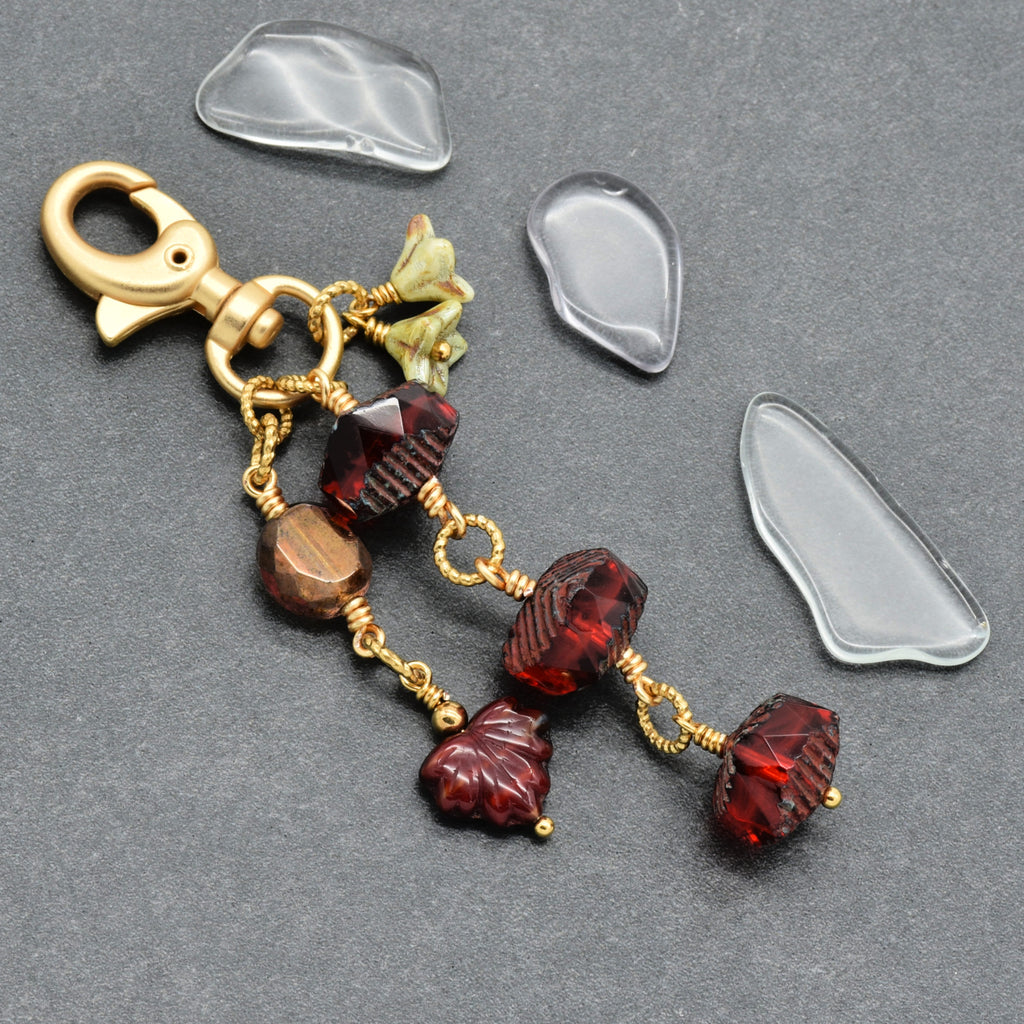 A handmade keychain featuring textured red and brown glass beads, gold wire-wrapped details, and a gold lobster clasp, arranged on a dark gray background.