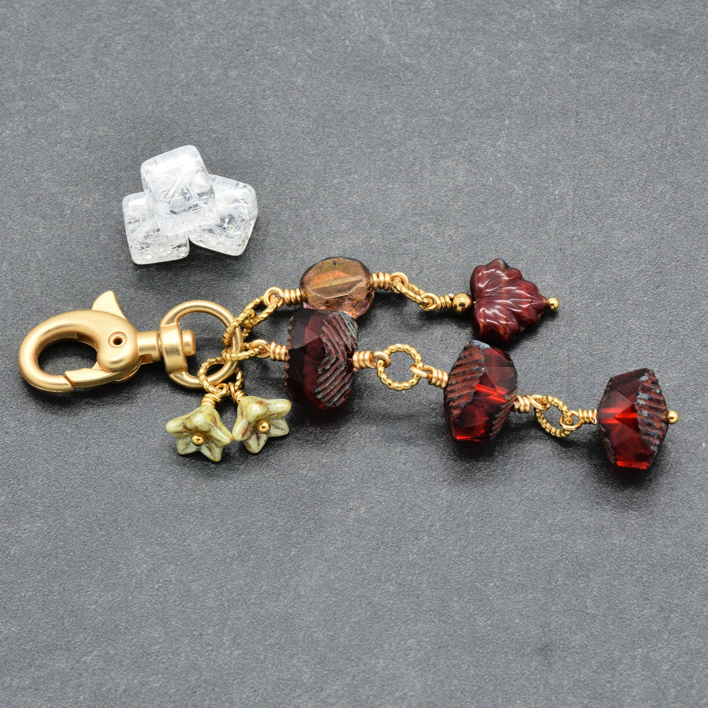 A handmade keychain charm featuring faceted dark red beads, small green flower charms, a smoky brown bead, and a gold clasp, displayed on a dark gray textured background next to a clear crystal.