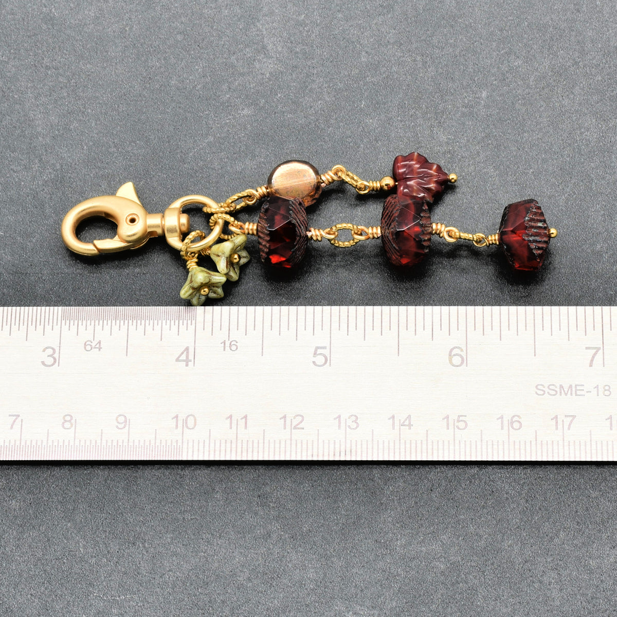 A close-up of a jewelry keychain or bag charm with a gold-tone lobster clasp, faceted dark red beads, small green flower charms, and a ruler below for scale, photographed on a dark gray textured background.
