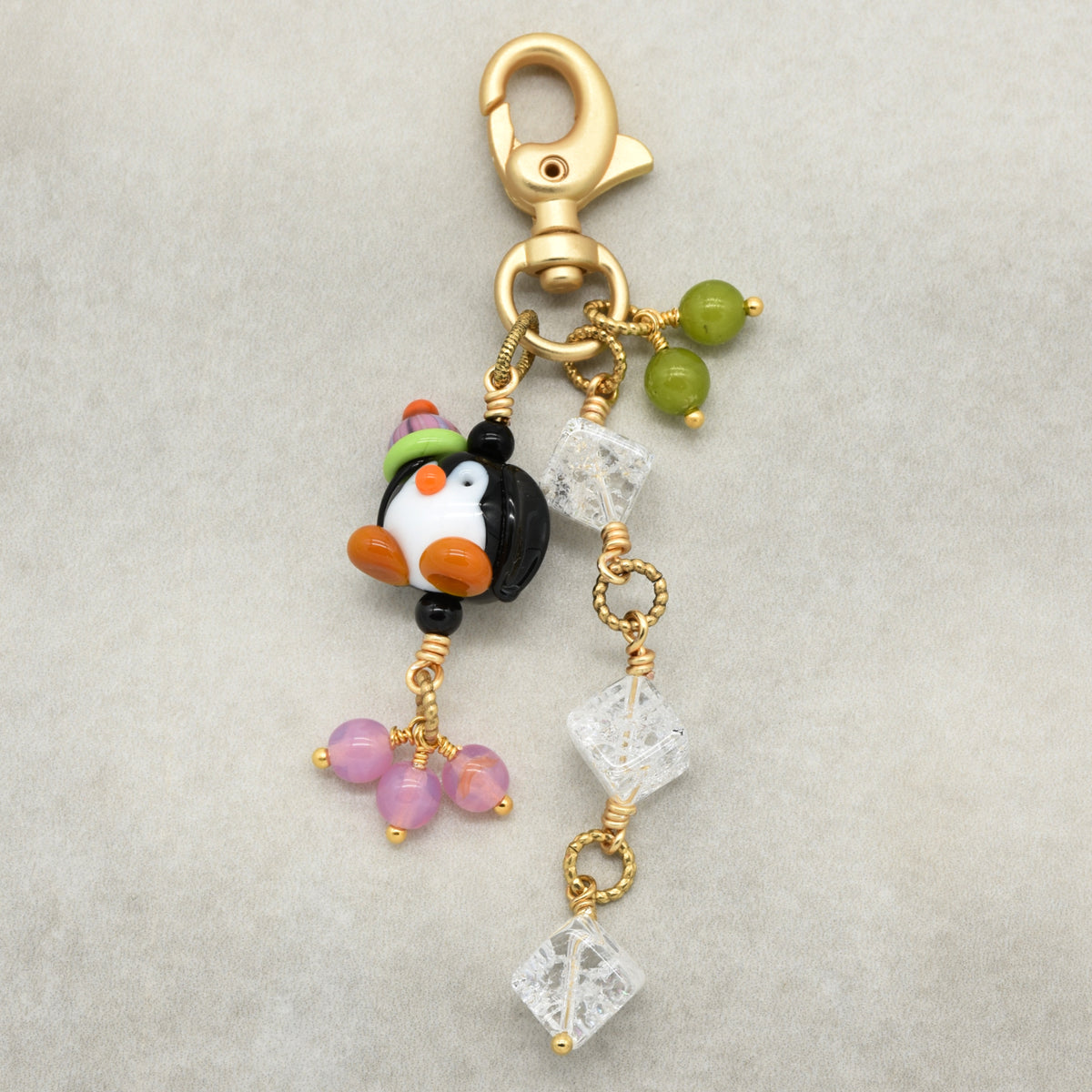 A gold-tone charm keychain featuring a small penguin bead with a green hat, clear quartz diamond-shaped beads, green and pink beads, and a gold lobster clasp, displayed on a neutral gray background.