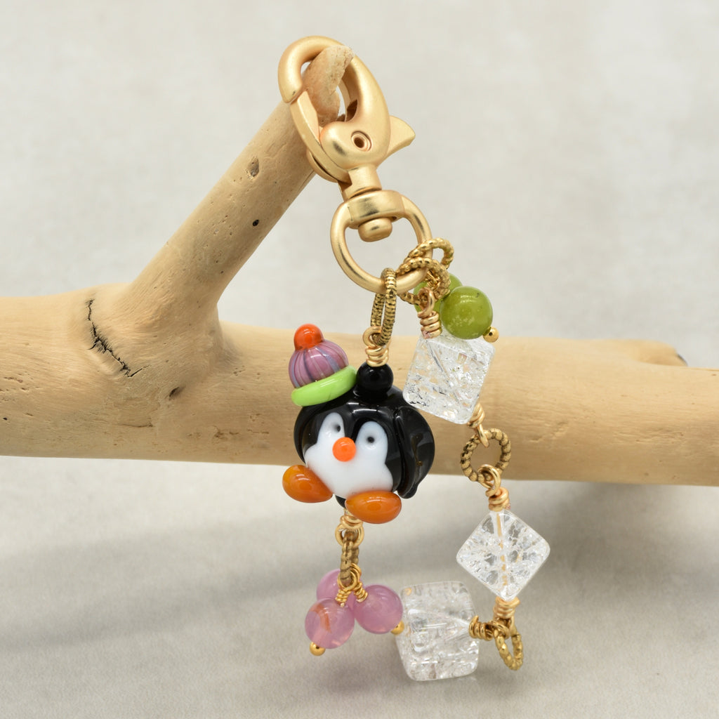 A gold-tone charm keychain featuring a small penguin wearing a purple and green hat, accented with clear faceted beads, pink and green beads, and a wooden branch prop against a neutral background.