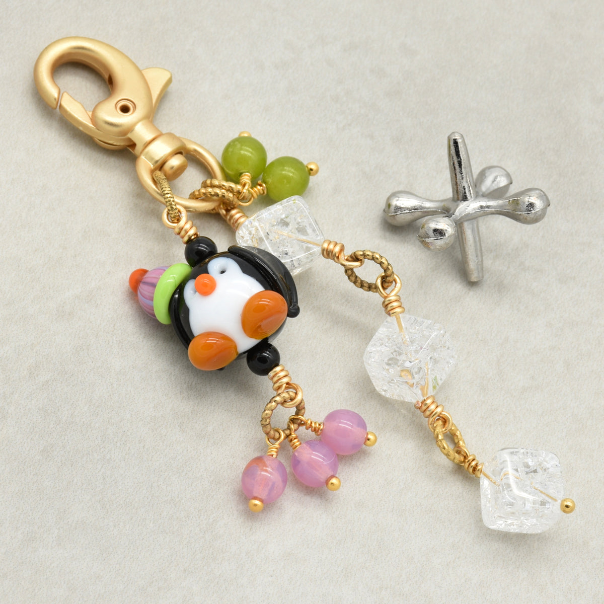 A handmade keychain charm featuring a black and white penguin bead with orange beak and feet, green beads, pink beads, clear crystal cube charms, and a gold lobster clasp. A separate silver metal charm is shown next to it on a light gray textured background.