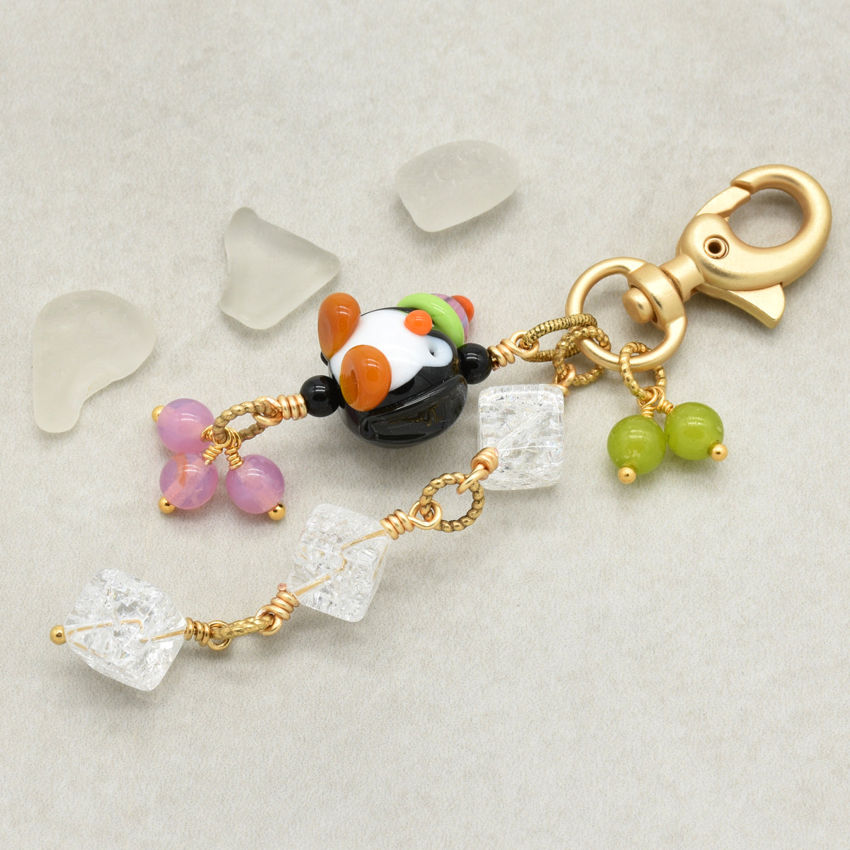 A handmade keychain charm featuring a small black and white penguin bead with orange and green accents, a gold lobster clasp, clear quartz crystal beads, pink beads, and green beads, displayed on a textured light gray background with decorative sea glass pieces.