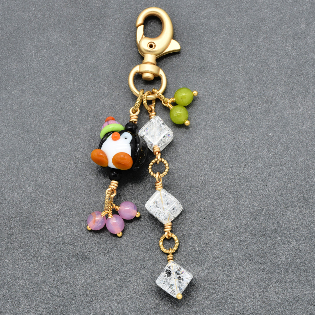 A gold-tone lobster clasp keychain featuring a small penguin charm with orange, green, and white details, alongside clear quartz crystal beads and small green and purple beads, displayed on a textured gray background.