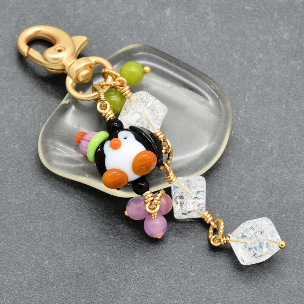 A handmade penguin charm keychain with a gold-tone lobster clasp, featuring a black and white penguin bead with orange beak and feet, green and pink beads, and clear quartz crystals, on a dark gray textured background.