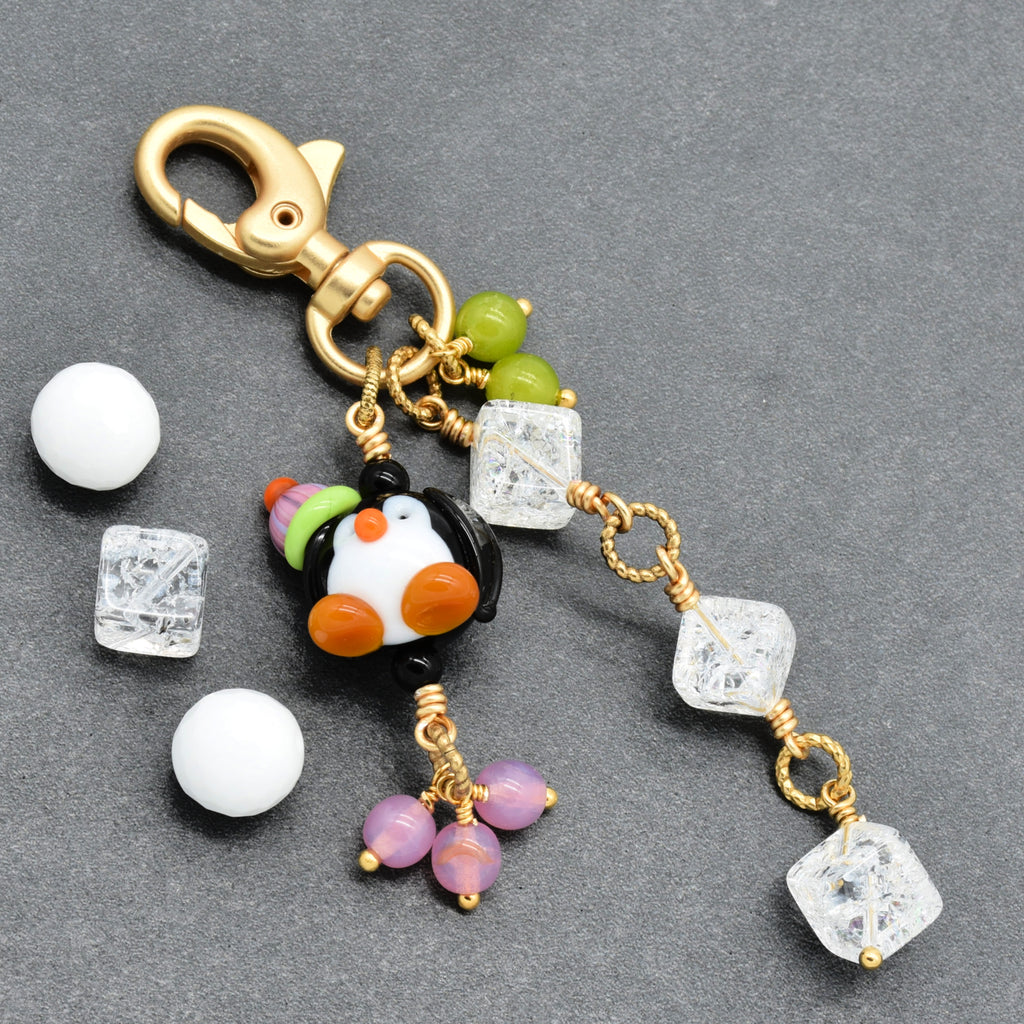 A gold-tone lobster clasp keychain charm featuring a small penguin bead, clear faceted crystal beads, purple beads, and green beads, displayed on a dark gray textured background.