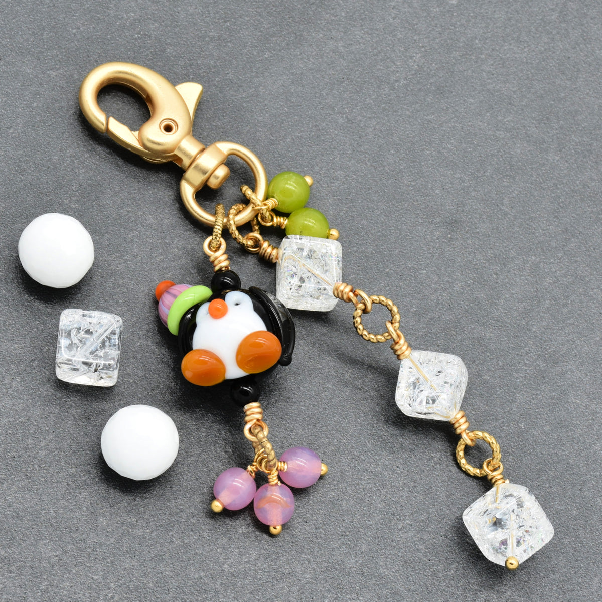 A gold-tone lobster clasp keychain charm featuring a small penguin bead, clear faceted crystal beads, purple beads, and green beads, displayed on a dark gray textured background.