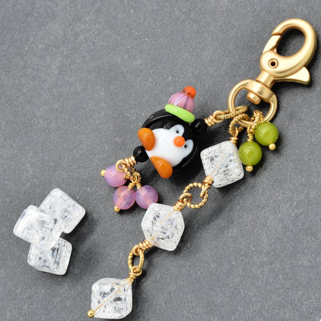 A decorative jewelry charm or keychain clasp with a penguin bead, gold-tone lobster clasp, green and purple beads, and clear faceted crystal-like drops on a dark gray background.