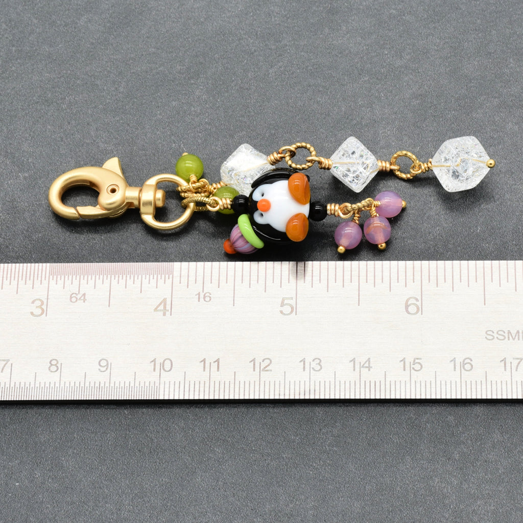 A handmade charm keychain with a gold clasp, featuring a penguin bead, clear crackle quartz beads, purple beads, and green beads, shown next to a metal ruler for scale on a dark gray background.