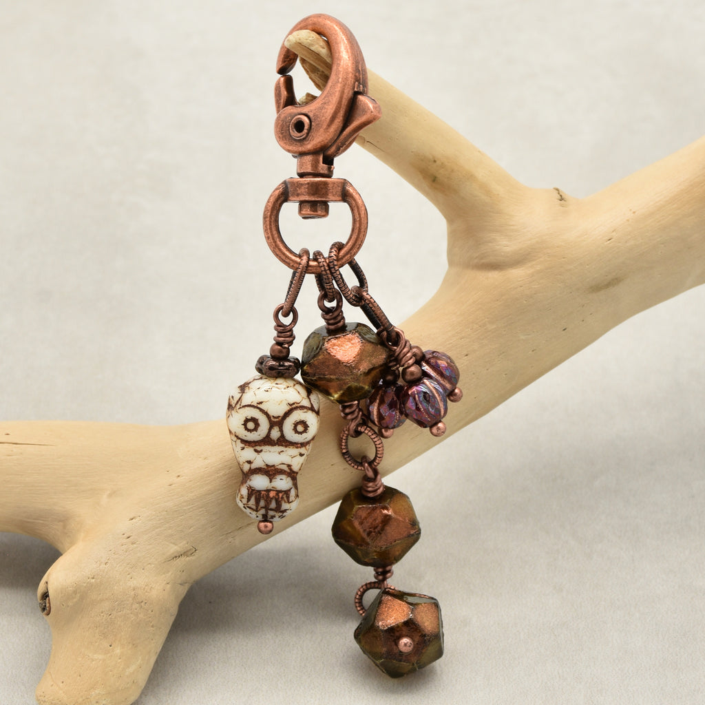 A handmade copper-toned keychain with an owl charm, faceted beads, and burgundy glass accents, displayed on a light wood branch against a neutral gray background - made with love in Colorado by Ruby Acorn.