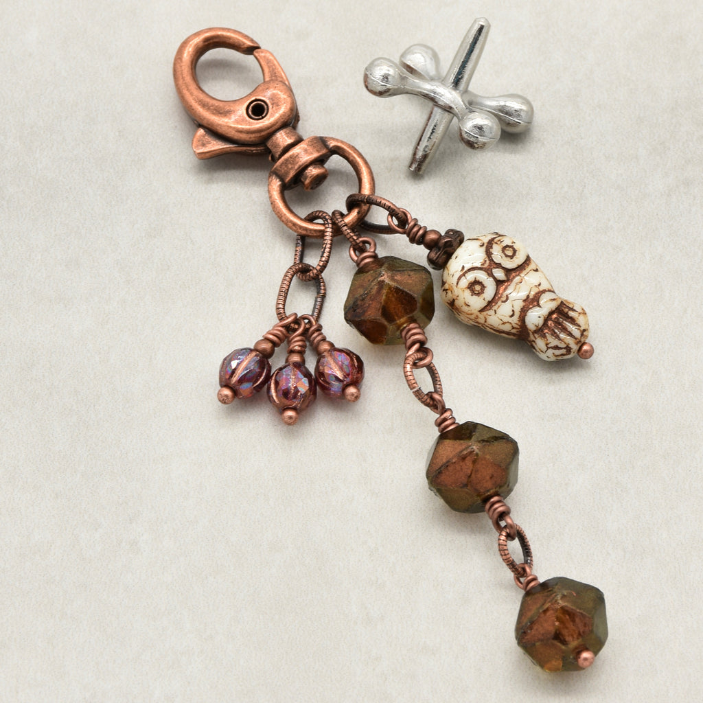 A handmade keychain or bag charm featuring a copper-toned lobster clasp, a an owl, faceted brown beads wrapped in copper wire, and three small iridescent burgundy beads, displayed on a light beige textured background - made with love in Colorado by Ruby Acorn.