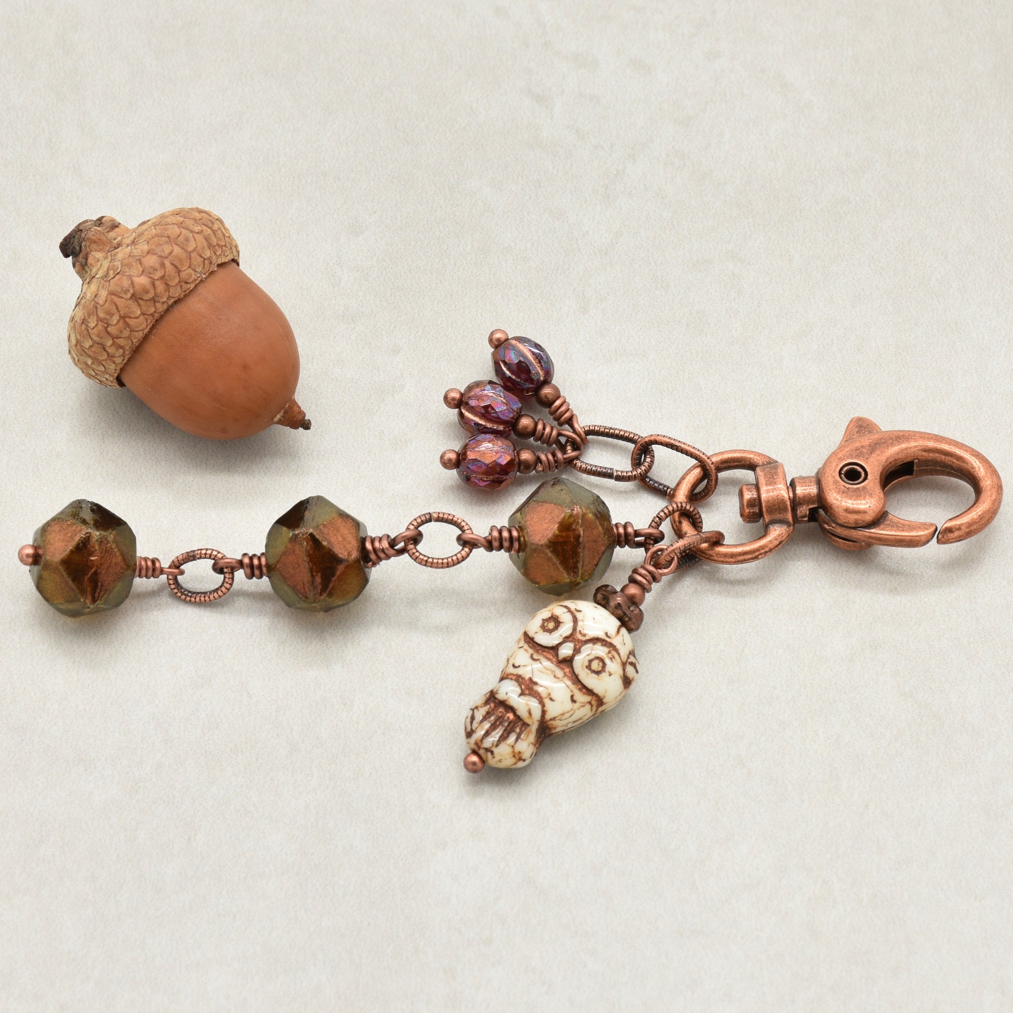 A handmade copper keychain featuring faceted bronze beads, a small owl bead, three burgundy fluted melons and a lobster clasp, displayed on a light beige textured background - made with love in Colorado by Ruby Acorn.